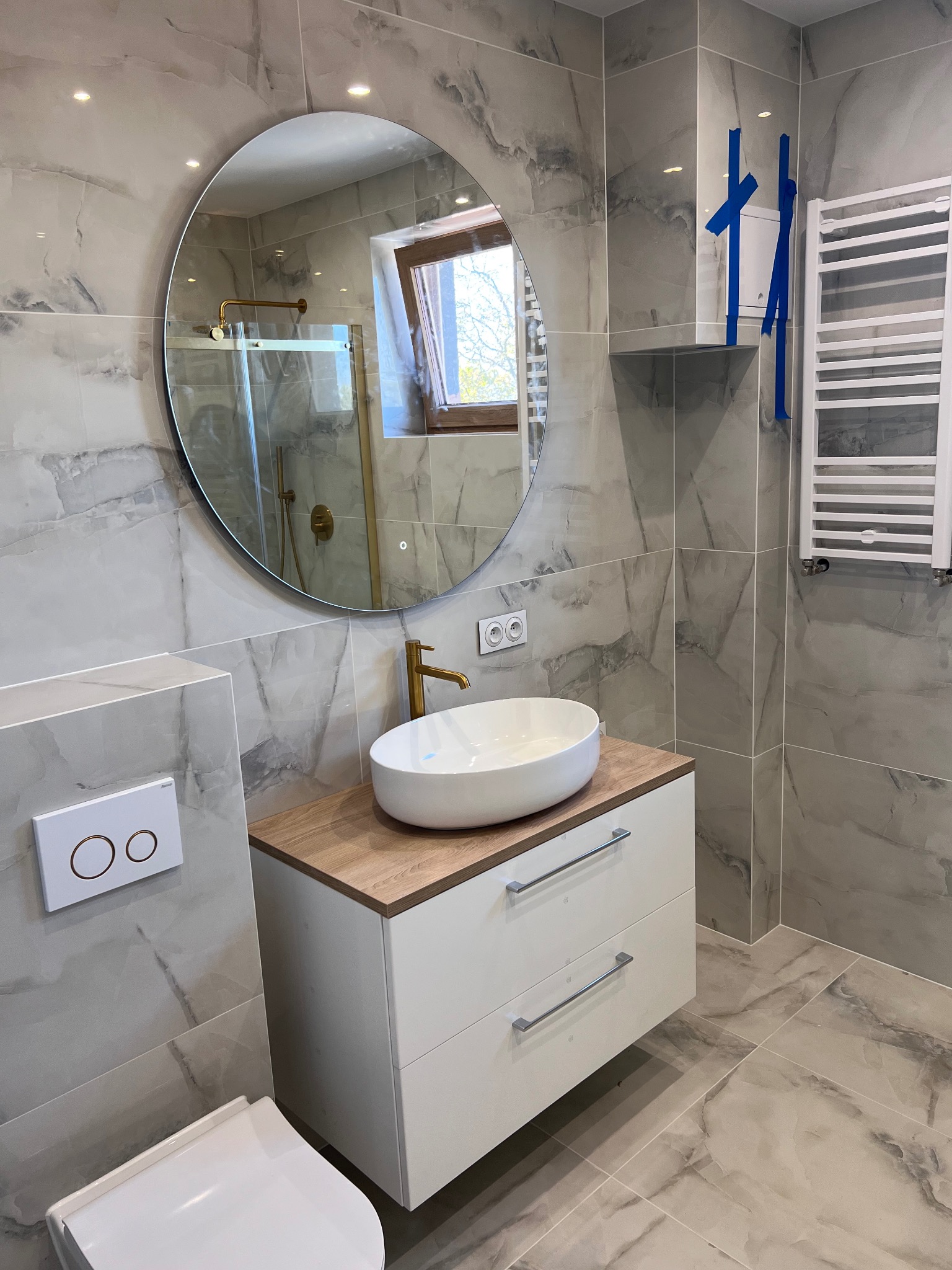 Nowoczesna łazienka z okrągłym lustrem, białą umywalką nablatową na szafce z drewnianym blatem i grzejnikiem drabinkowym. Szare płytki imitujące marmur na ścianach i podłodze.