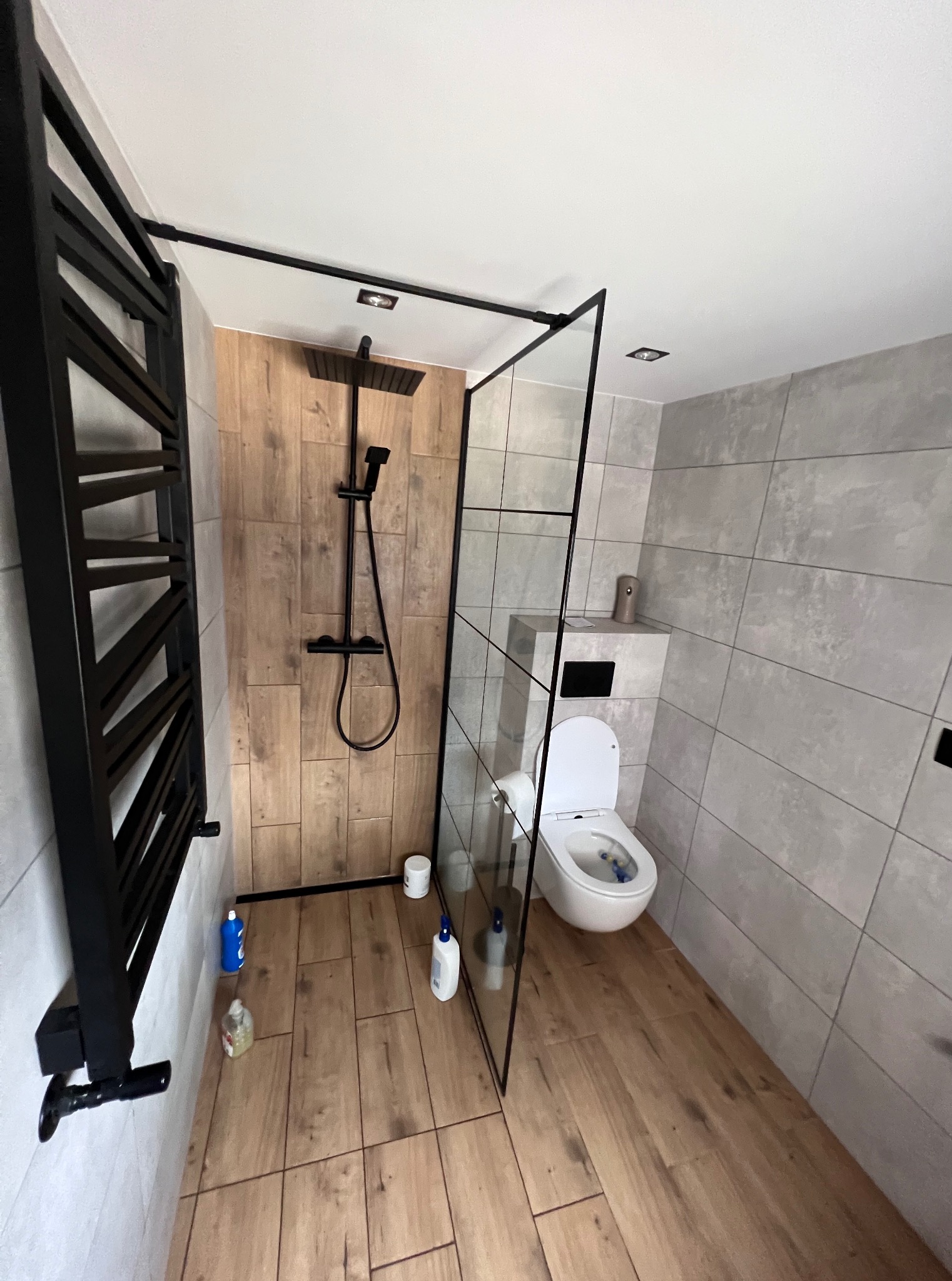 Nowoczesna łazienka po remoncie: kabina walk-in z czarnymi profilami, grzejnik drabinkowy, płytki imitujące drewno na podłodze i pod prysznicem, szare płytki na ścianach.