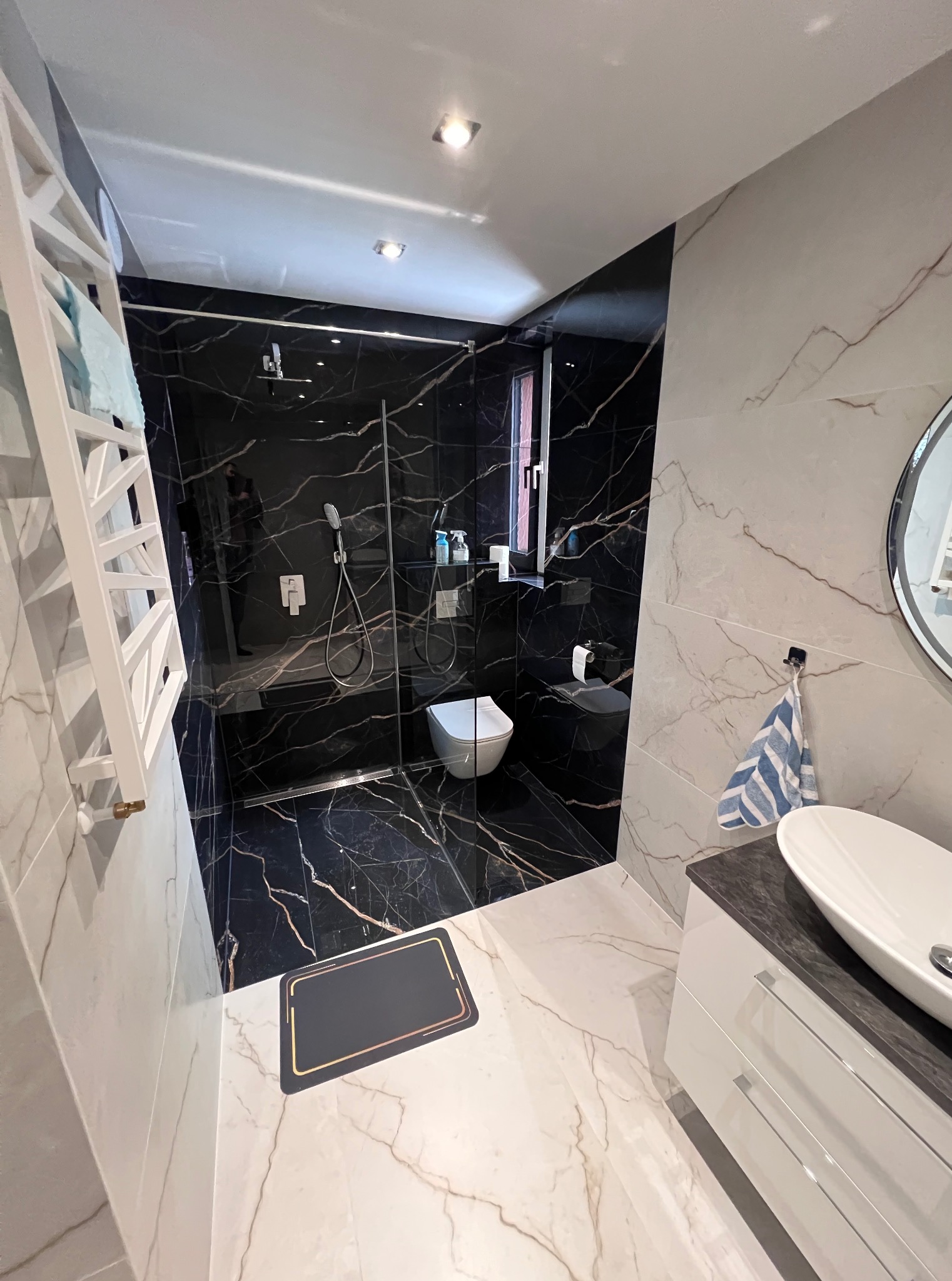 Nowoczesna łazienka z kabiną typu walk-in, czarne i białe marmurowe płytki, biała umywalka nablatowa i grzejnik drabinkowy, akcenty kolorystyczne w postaci ręczników.