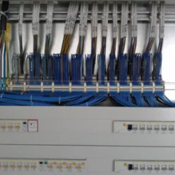 Szczegółowe ujęcie rozdzielni elektrycznej z licznymi niebieskimi przewodami, bezpiecznikami i oznaczeniami, ukazujące profesjonalnie wykonaną instalację.