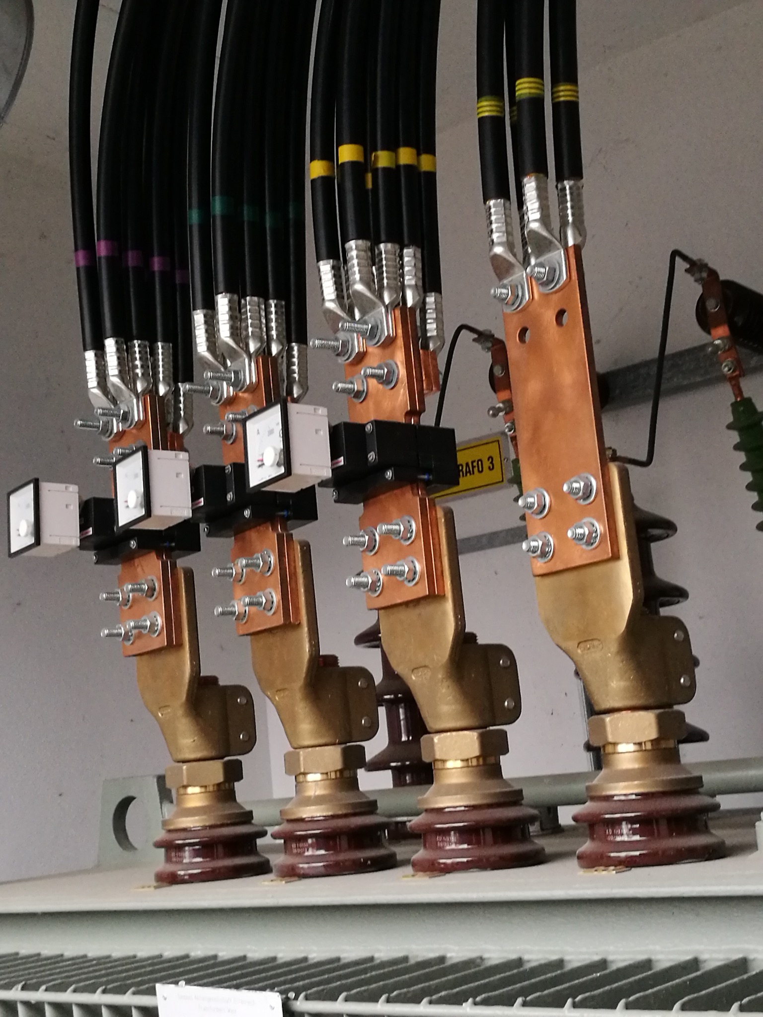 Szczegółowe ujęcie transformatora z podłączonymi przewodami i miernikami, ukazujące precyzję instalacji elektrycznej. Widoczne oznaczenia kolorystyczne na przewodach.