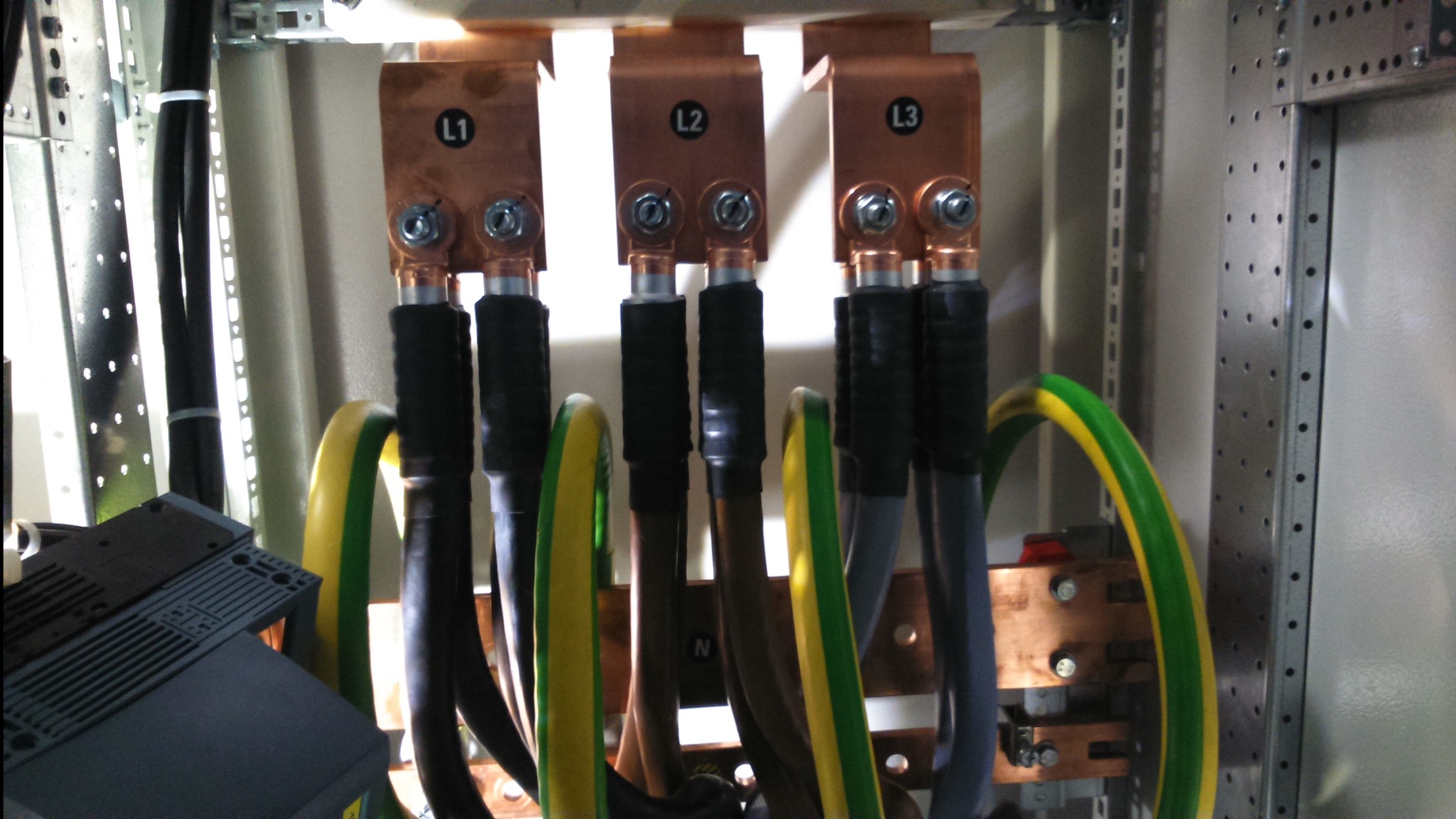 Szczegółowe ujęcie rozdzielnicy elektrycznej z miedzianymi szynami oznaczonymi L1, L2, L3 i N, z podłączonymi przewodami w kolorach czarnym, szarym i żółto-zielonym.