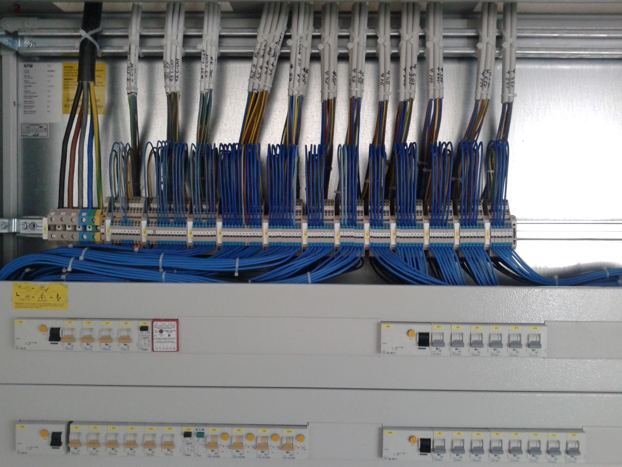 Szczegółowe ujęcie rozdzielni elektrycznej z licznymi niebieskimi przewodami, bezpiecznikami i oznaczeniami, ukazujące profesjonalnie wykonaną instalację.