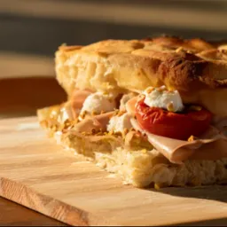 Przekrojona focaccia z szynką, pomidorem i serem, posypana pistacjami, leżąca na drewnianej desce w ciepłym świetle. Widoczne detale struktury pieczywa i świeżość składników.
