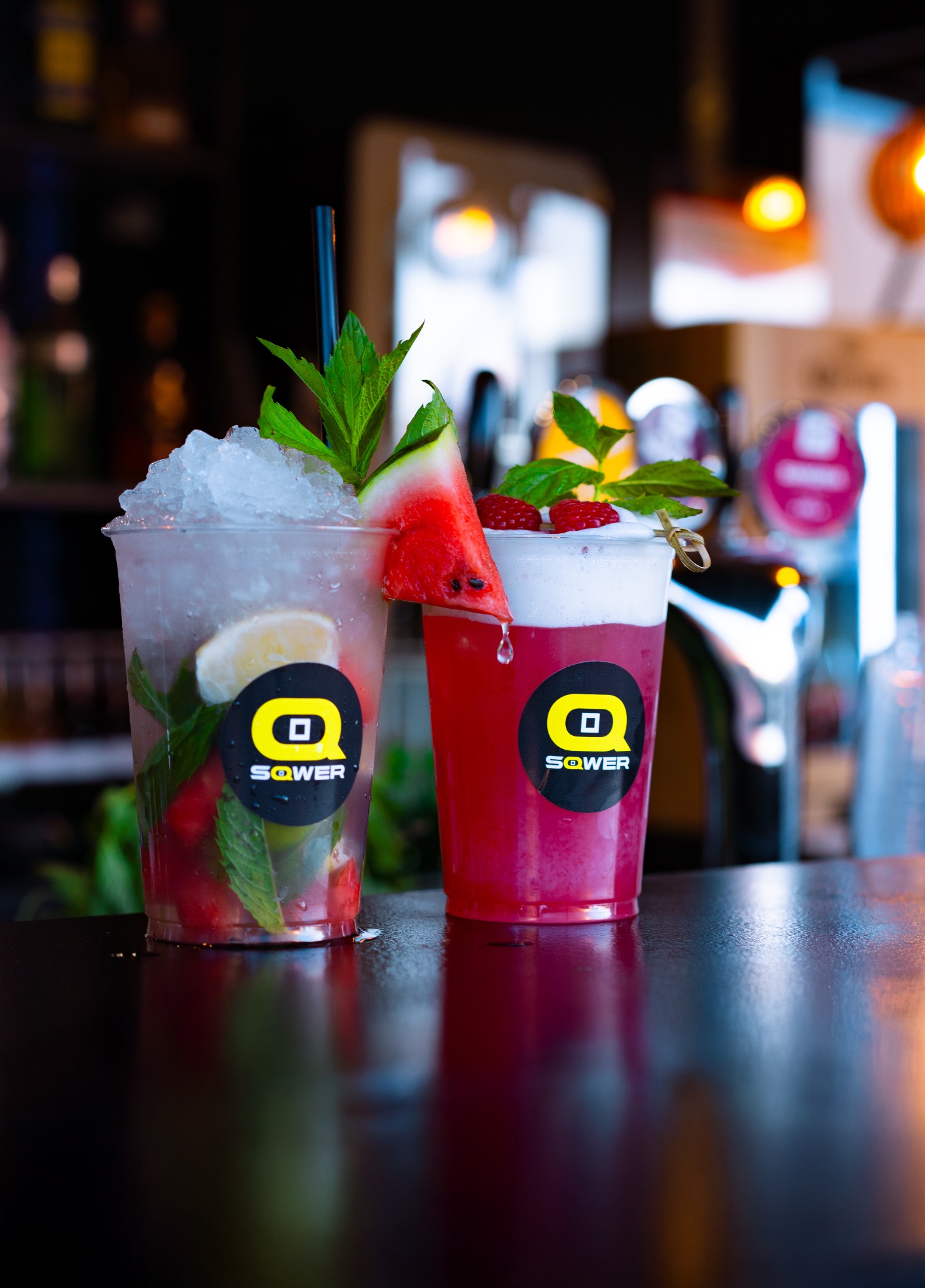 Dwa kolorowe drinki w plastikowych kubkach z logiem, udekorowane owocami i miętą, stojące na barze. Jeden z lodem, drugi z pianką. W tle barowe wyposażenie.