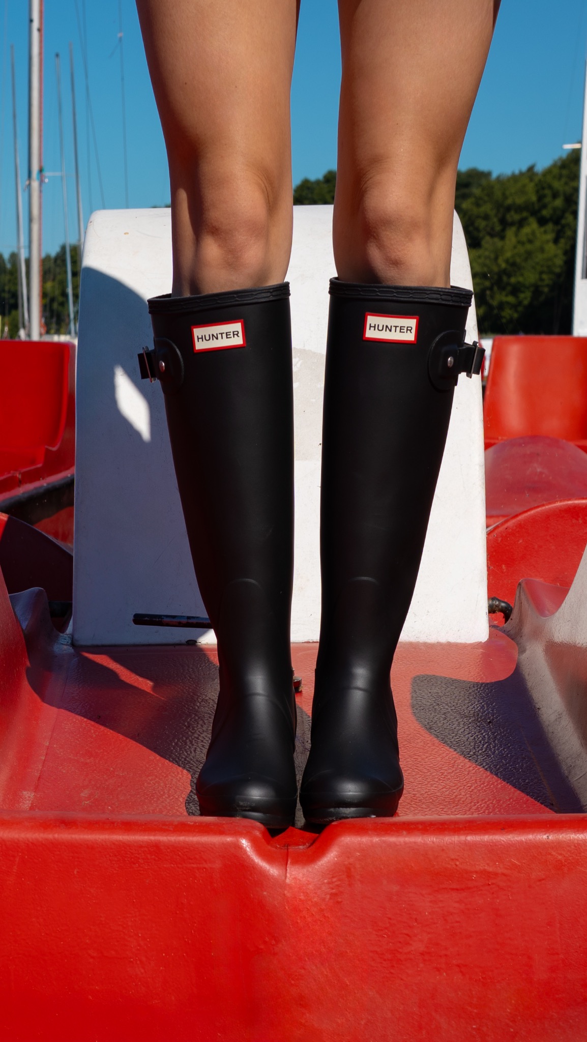 Osoba w czarnych kaloszach Hunter stoi na czerwonym rowerze wodnym, w tle błękitne niebo i zieleń. Ujęcie od kolan w dół, w słoneczny dzień.