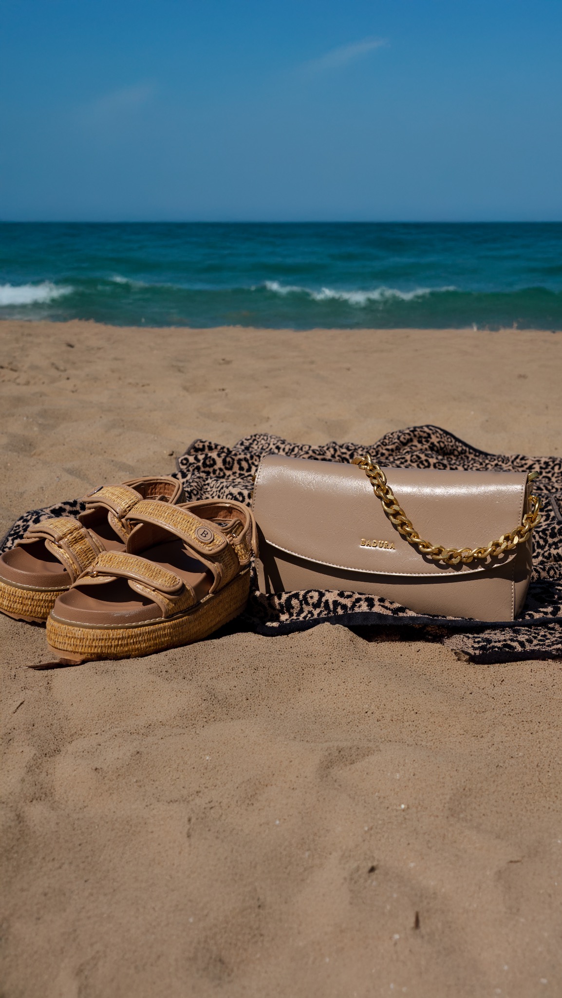 Beżowa torebka z łańcuchem i sandały na platformie leżą na kocu w panterkę na piaszczystej plaży z widokiem na morze i błękitne niebo w tle.