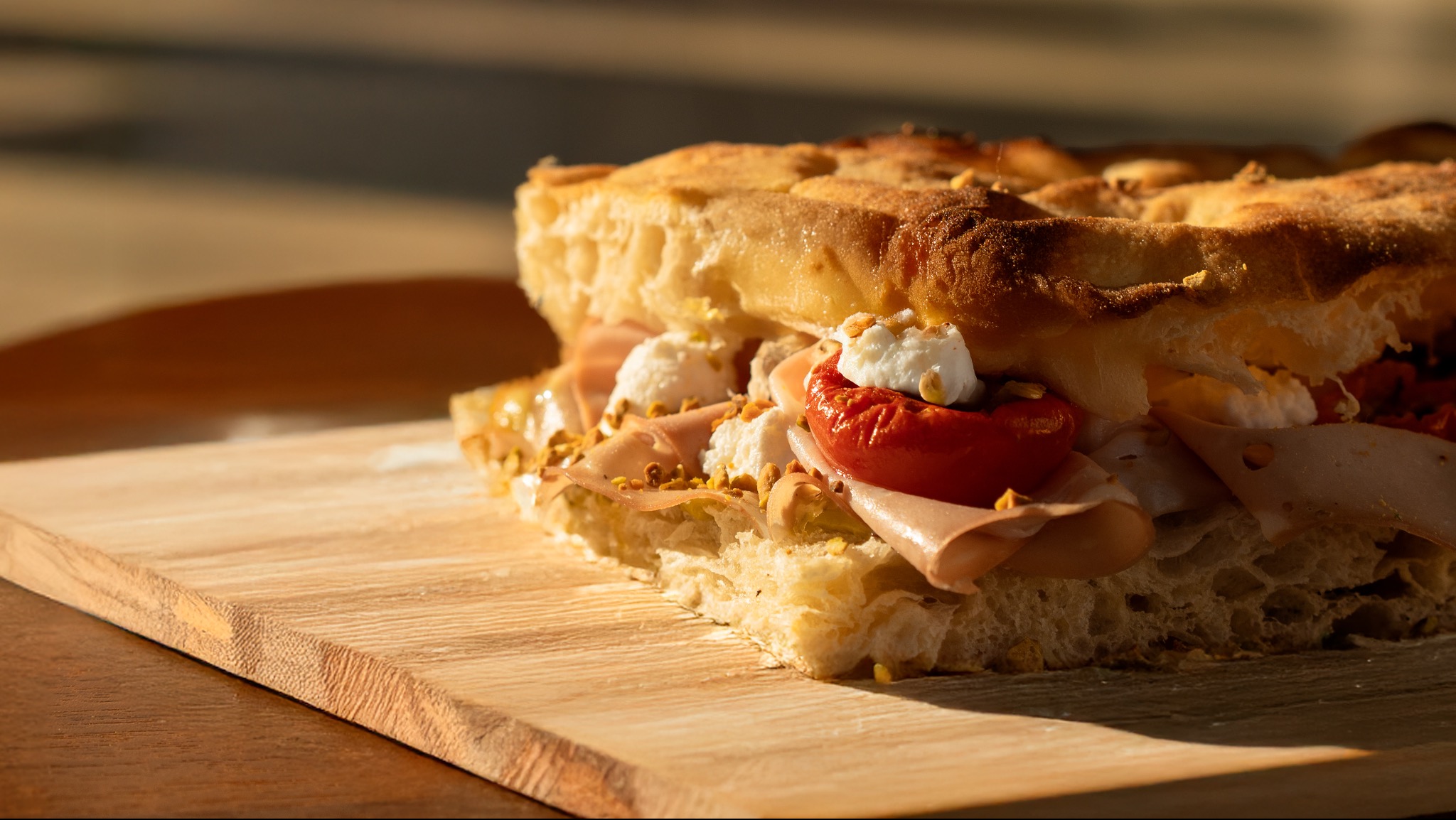 Przekrojona focaccia z szynką, pomidorem i serem, posypana pistacjami, leżąca na drewnianej desce w ciepłym świetle. Widoczne detale struktury pieczywa i świeżość składników.