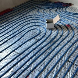 Daniel Łapiński - Instalacja ogrzewania podłogowego w trakcie układania: niebieskie rurki na srebrnej folii, czarna siatka, białe pudełko z akcesoriami w rogu pomieszczenia.