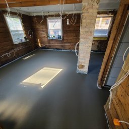 Daniel Łapiński - Wylany, gładki betonowy podkład podłogowy w remontowanym pomieszczeniu z drewnianymi ścianami i oknami. Widoczny fragment ceglanego filaru i wystające przewody.