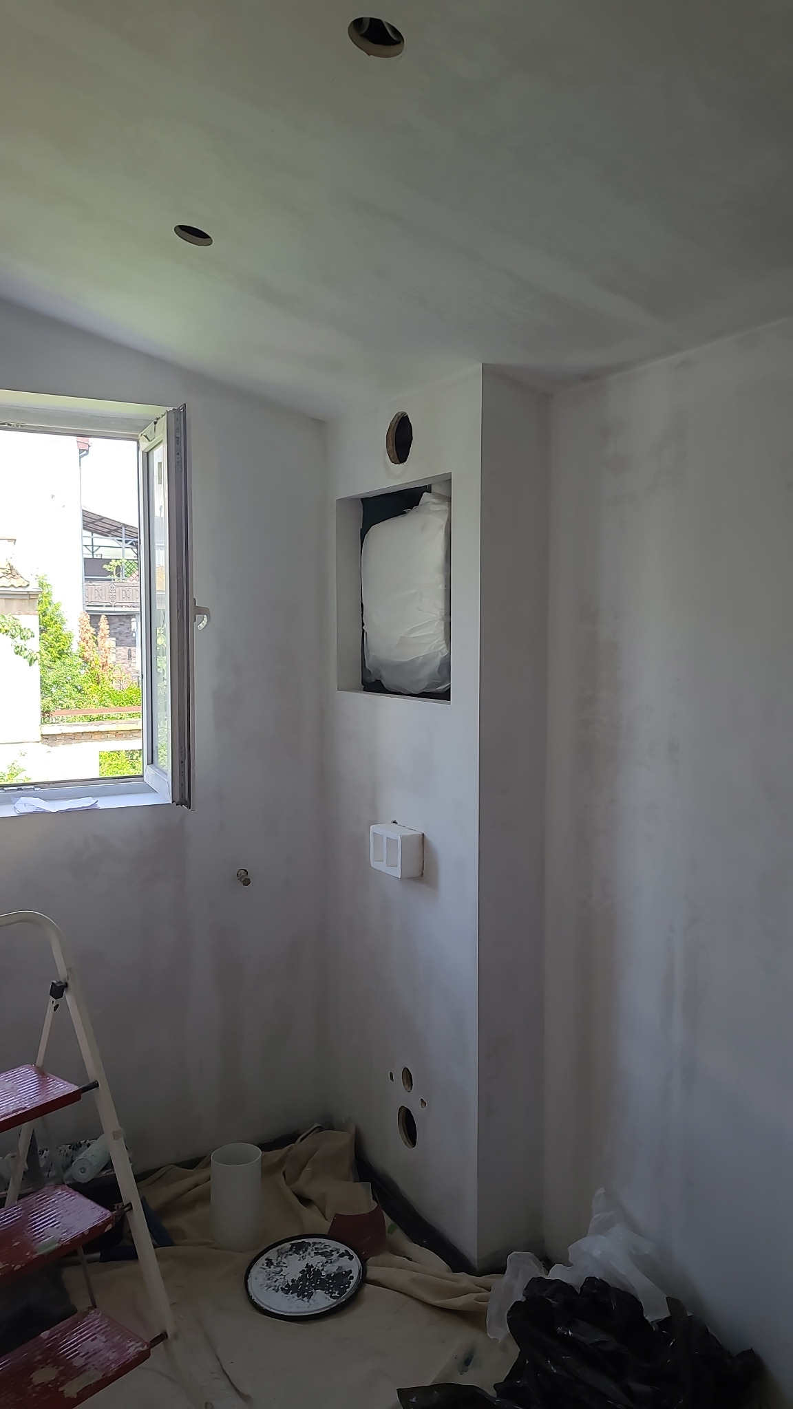 Wnętrze pokoju po szpachlowaniu ścian, widoczne otwory w suficie, okno z widokiem na zieleń, drabina i akcesoria malarskie. Ściany w jasnym odcieniu.