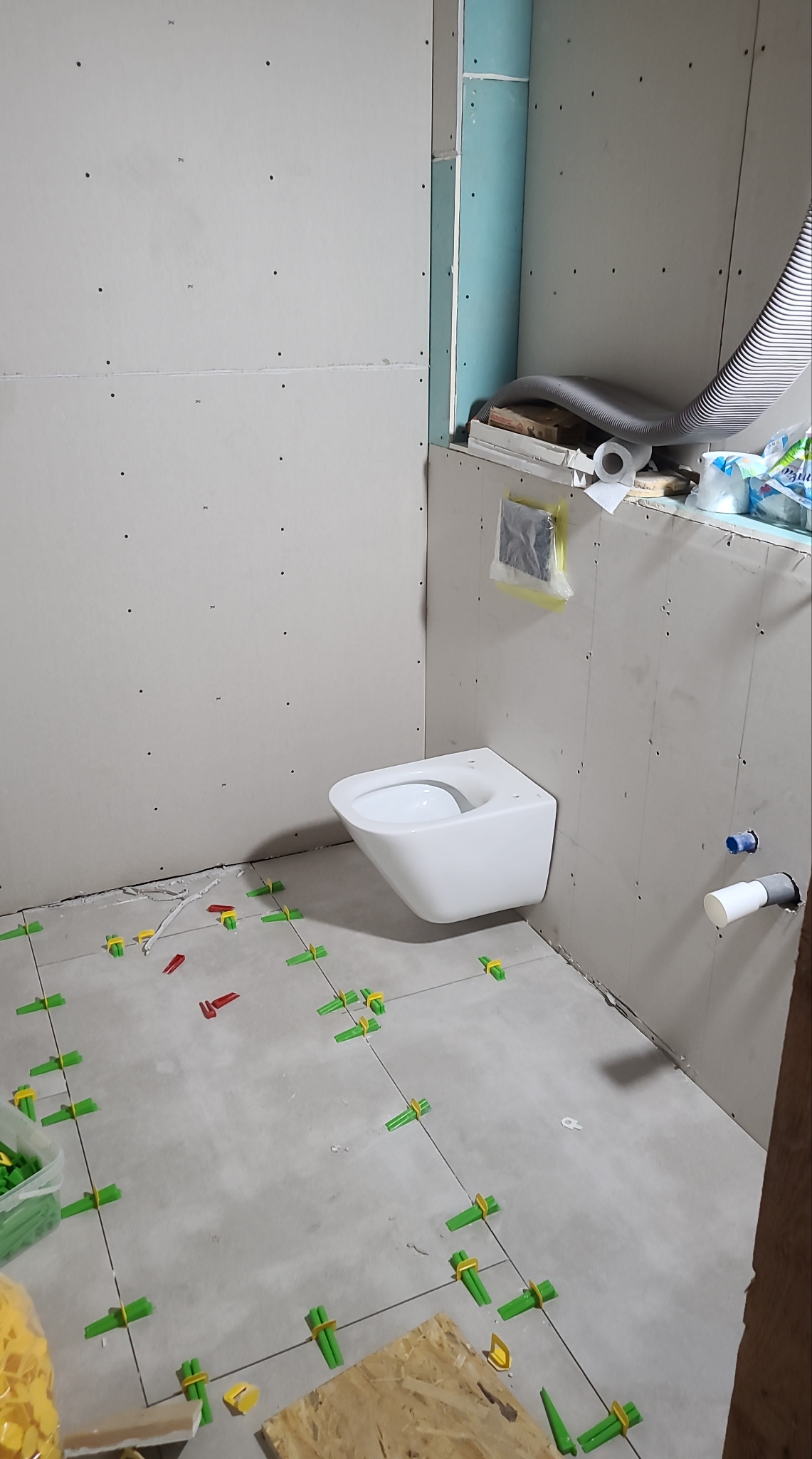 Łazienka w trakcie wykańczania: biała wisząca toaleta, szare płytki podłogowe z zielonymi krzyżykami, ściany z płyt gipsowo-kartonowych przed wykończeniem.