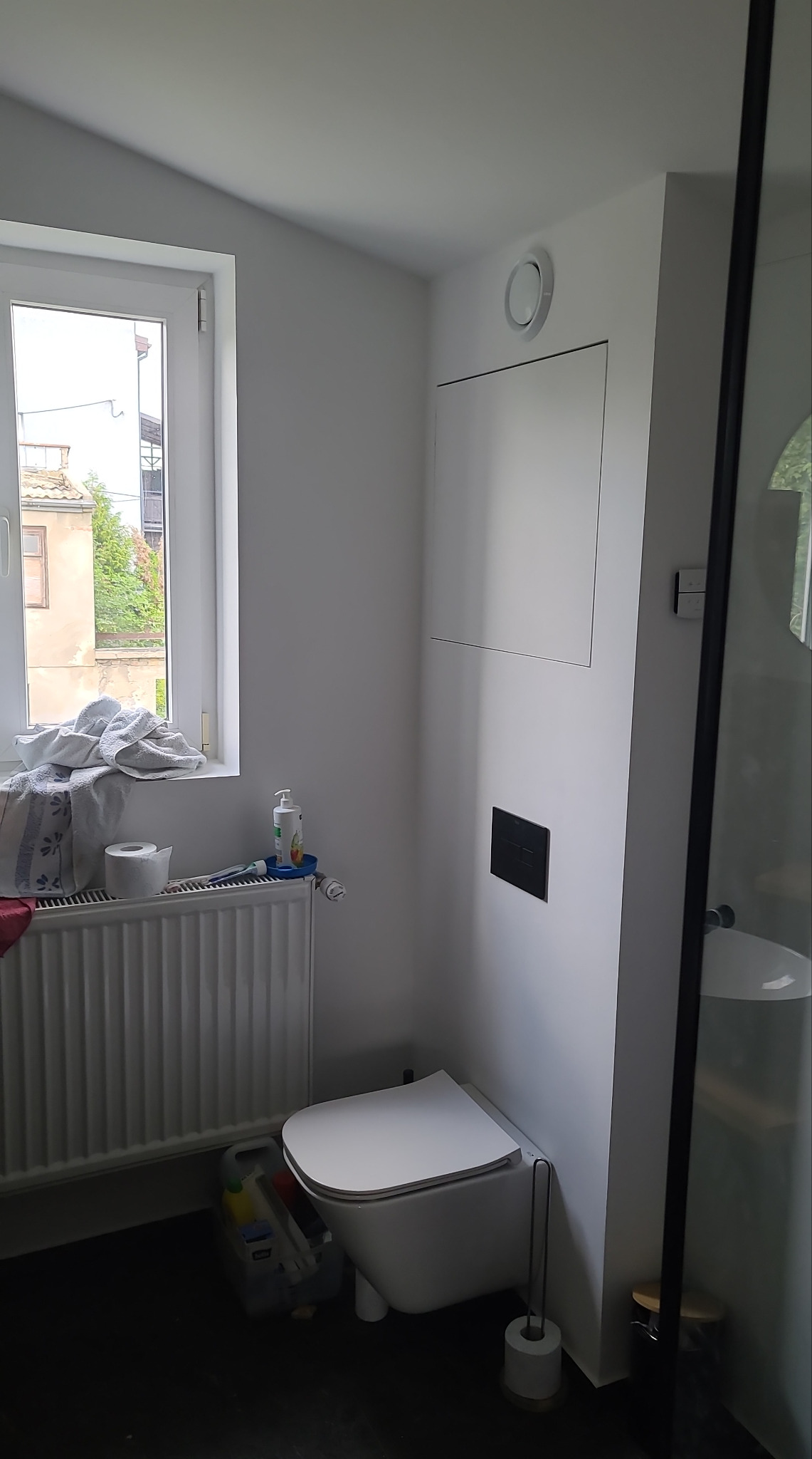 Nowoczesna, minimalistyczna łazienka z podwieszaną toaletą, grzejnikiem pod oknem i czarną armaturą. Widoczne okno z widokiem na budynek.