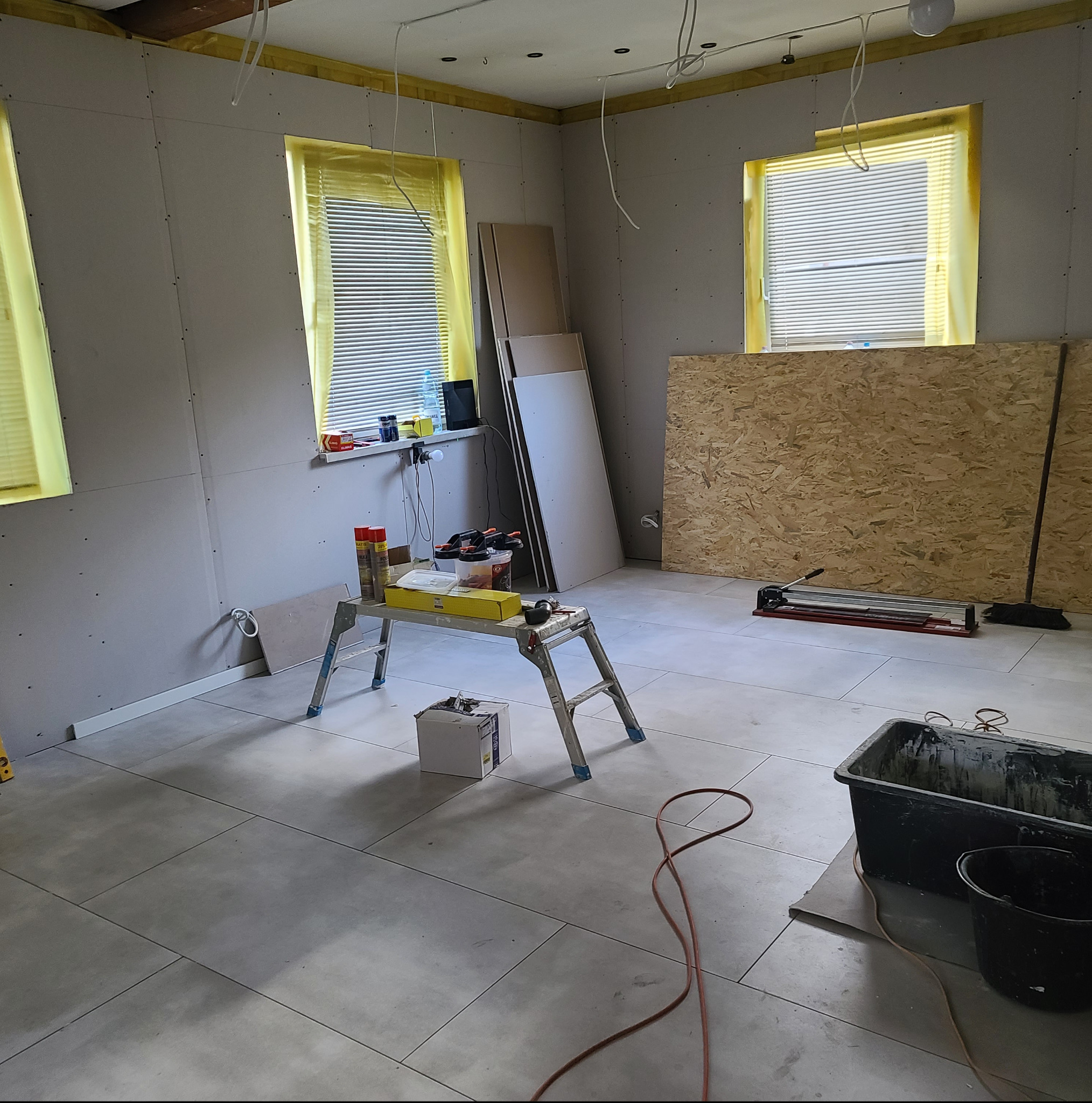 Wnętrze w trakcie prac: położone płytki podłogowe, ściany z płyt gipsowych, okna zabezpieczone folią, narzędzia na podeście. Widoczne płyty i akcesoria glazurnicze.