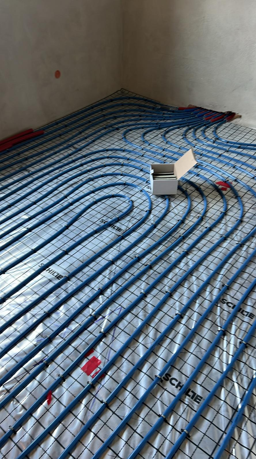 Instalacja ogrzewania podłogowego w trakcie układania: niebieskie rurki na srebrnej folii, czarna siatka, białe pudełko z akcesoriami w rogu pomieszczenia.