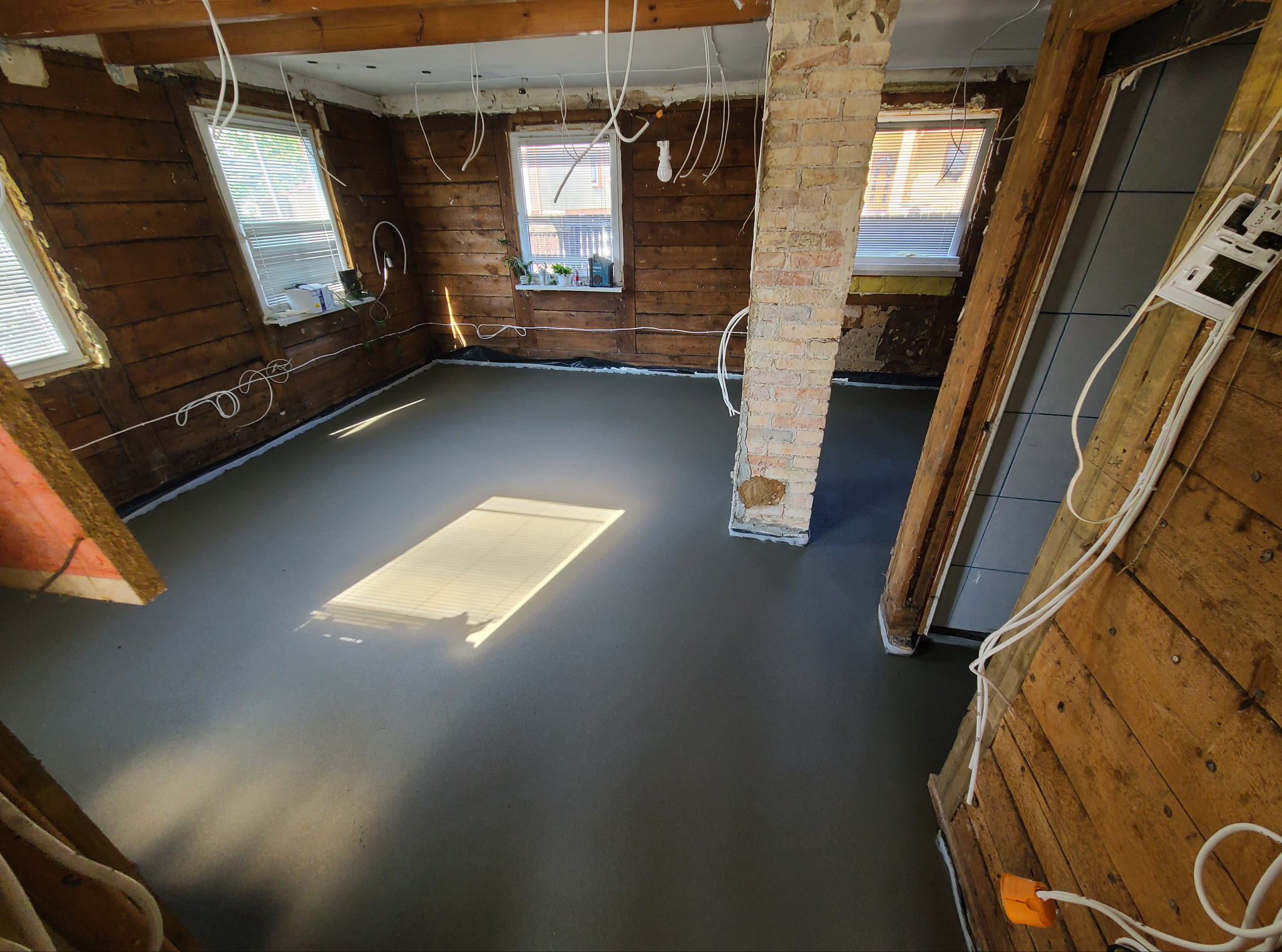 Wylany, gładki betonowy podkład podłogowy w remontowanym pomieszczeniu z drewnianymi ścianami i oknami. Widoczny fragment ceglanego filaru i wystające przewody.