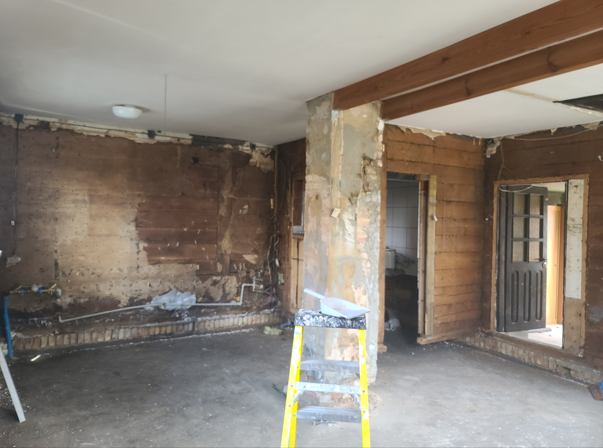 Wnętrze w trakcie demontażu: odsłonięte drewniane ściany, fragmenty tynku, drabina malarska z narzędziami. Widoczne instalacje i przygotowania do dalszych prac.