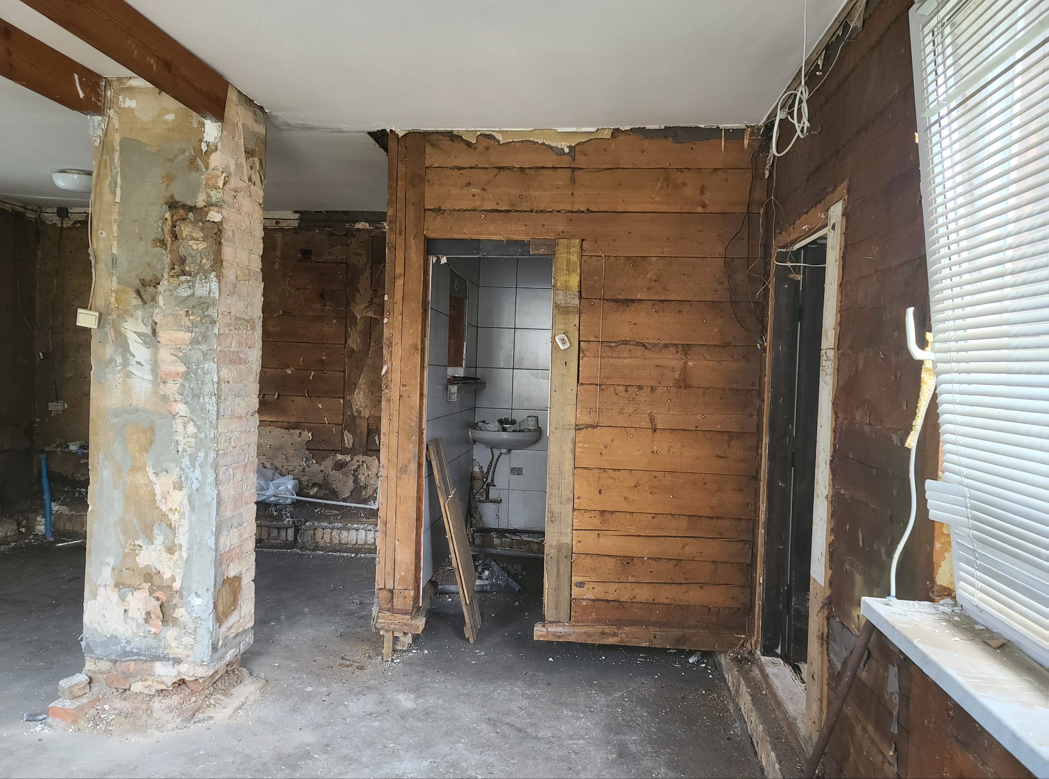 Wnętrze w trakcie remontu: odsłonięte cegły, drewniane ściany, widoczne instalacje. Fragment łazienki z umywalką w tle. Okno z żaluzjami po prawej.