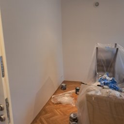 CONTRAST GRZEGORZ BORKOWSKI - Świeżo pomalowany pokój z parkietem, zabezpieczone meble folią, pędzle i farby. Widoczne detale wykończenia sufitu. Jasne, naturalne światło.