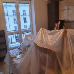 CONTRAST GRZEGORZ BORKOWSKI - Przygotowanie do prac malarskich: meble i podłoga zabezpieczone folią malarską w jasnym wnętrzu z oknem na kamienicę. Widoczne narzędzia malarskie.