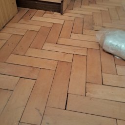 Parkiet ułożony w jodełkę, widoczne szczeliny i drobne zabrudzenia. Fragment mebla w lewym górnym rogu. Częściowo widoczna folia ochronna na podłodze.