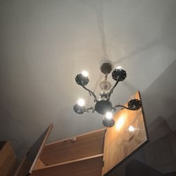 Antyczny żyrandol z żarówkami, widoczny z dołu, oświetla otwartą szafę z drewnianymi półkami. Nietypowy kadr.