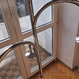 Fragment balkonu z parkietem w jodełkę i metalową barierką, widok z góry. Okno balkonowe z widokiem na zewnątrz. Częściowo widoczna ściana z nierówną fakturą.