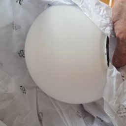 Biała, okrągła lampa w papierowym opakowaniu, częściowo wyjęta z pudełka. Widoczna dłoń osoby wyjmującej lampę. Delikatne światło podkreśla gładką powierzchnię.