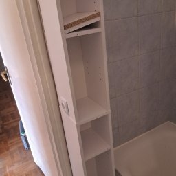 CONTRAST GRZEGORZ BORKOWSKI - Wąska, biała szafka łazienkowa z półkami, częściowo zamontowana, widoczna obok wanny i ściany z szarych płytek. Ujęcie z bliska, z widocznym gniazdkiem elektrycznym.