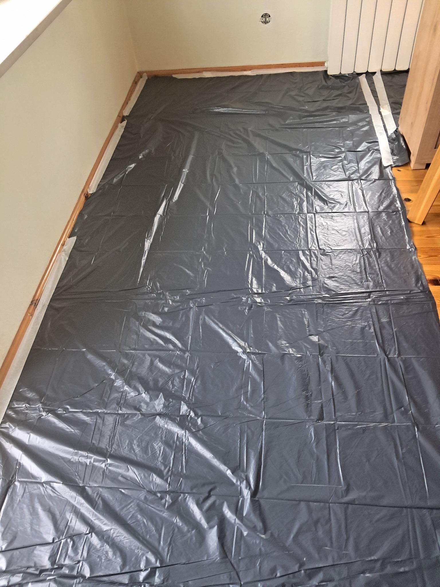 Zabezpieczona folią podłoga w pokoju przed pracami remontowymi. Widoczne listwy przypodłogowe i fragment grzejnika. Przygotowanie do malowania lub innego etapu wykończenia.