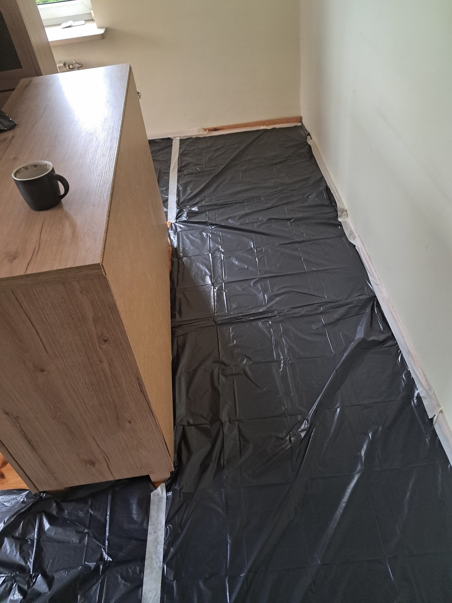 Przygotowanie do malowania: podłoga zabezpieczona folią i taśmą malarską, meble odsunięte, kubek na szafce. Widok z góry, wnętrze w trakcie prac.