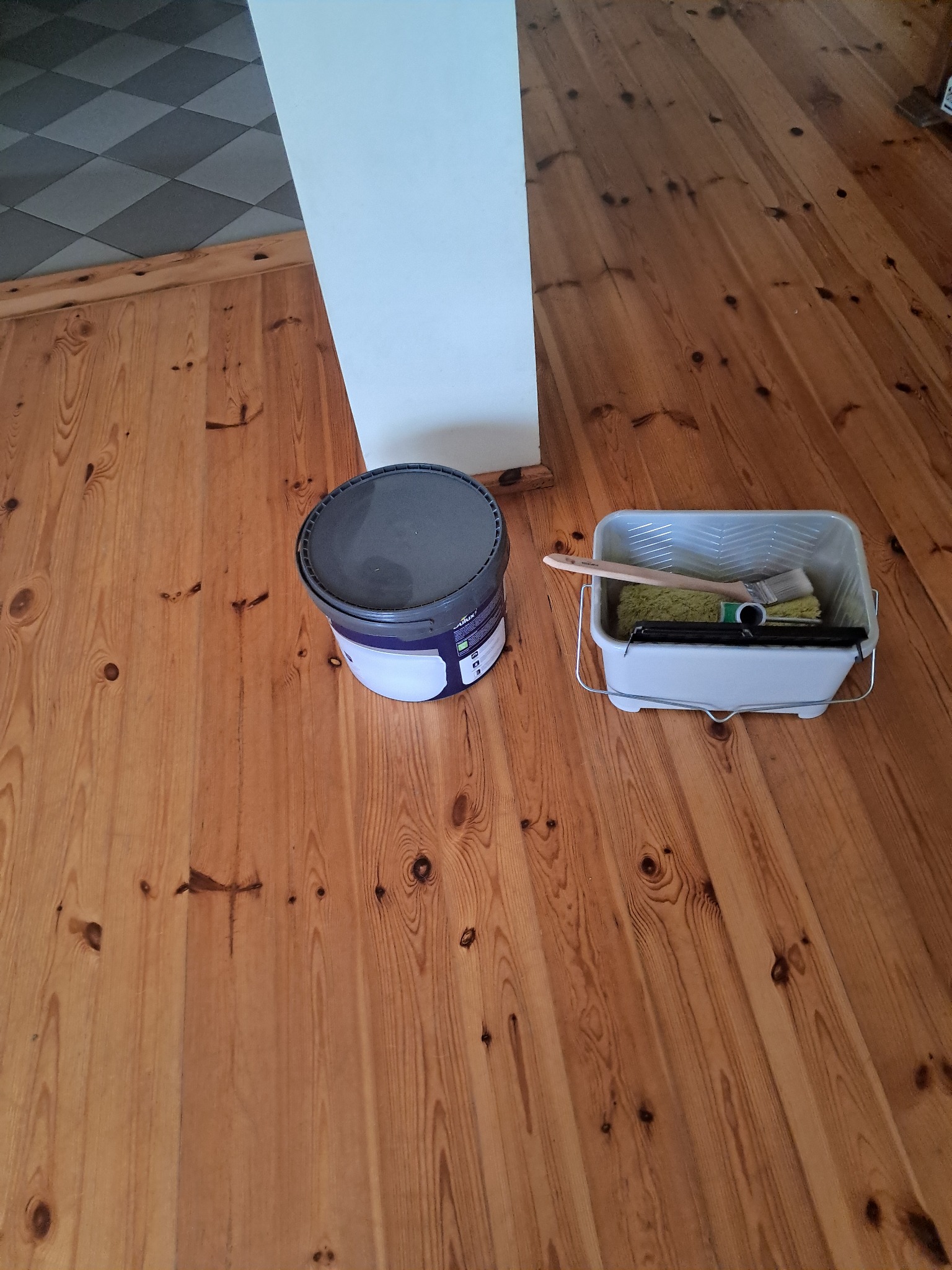 Przygotowanie do malowania podłogi: wiadro z farbą, kuweta malarska z wałkiem i pędzlem na drewnianej podłodze w odcieniach brązu, przy białej ścianie i szarej podłodze.