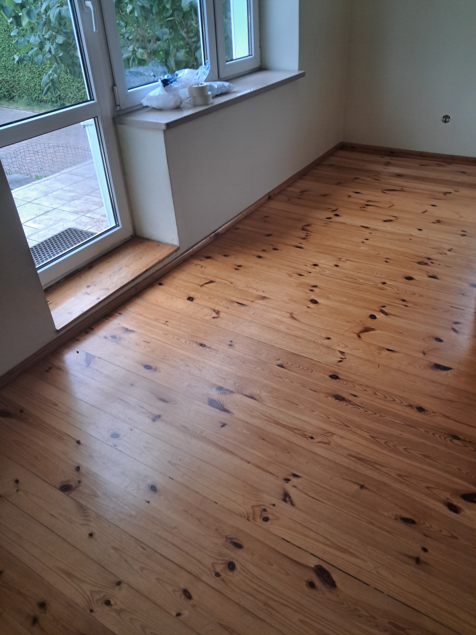 Wykończona podłoga z desek w odcieniach brązu, widoczna z perspektywy narożnika pokoju z oknem. Świeżo pomalowane ściany i parapet. Widoczne ślady po taśmie malarskiej.