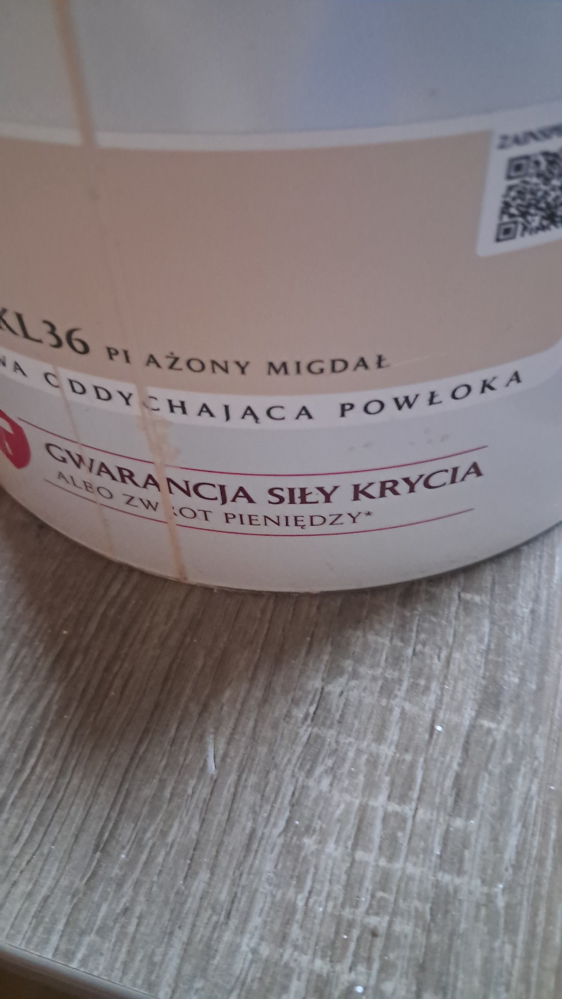 Puszka farby w odcieniu 'Piażony Migdał' z napisem 'Gwarancja siły krycia albo zwrot pieniędzy' na tle podłogi imitującej drewno.