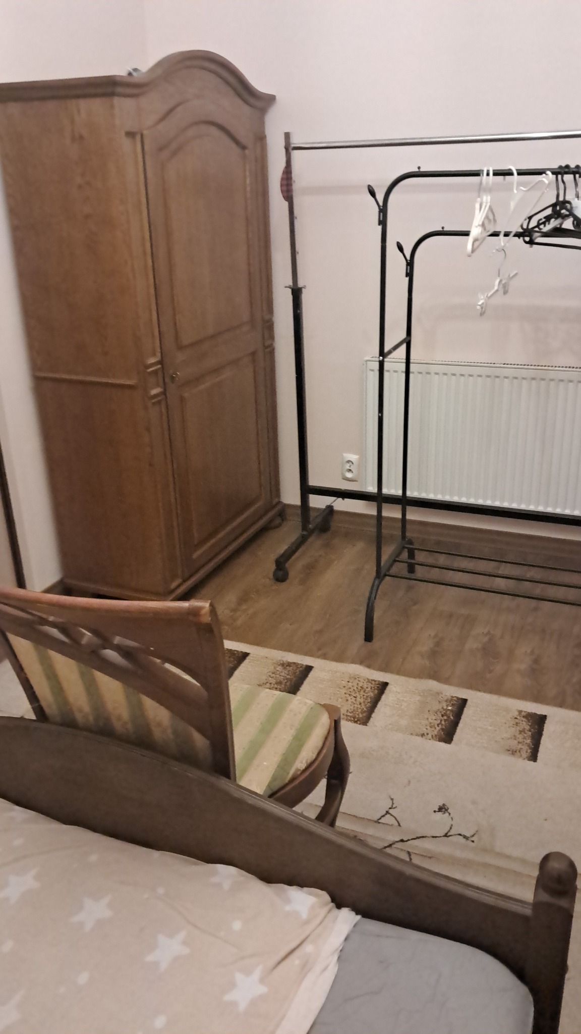 Drewniana szafa z dwojgiem drzwi, w tle wieszak na ubrania i grzejnik. Widok z perspektywy łóżka z drewnianym wezgłowiem i krzesłem z zielonym siedziskiem.