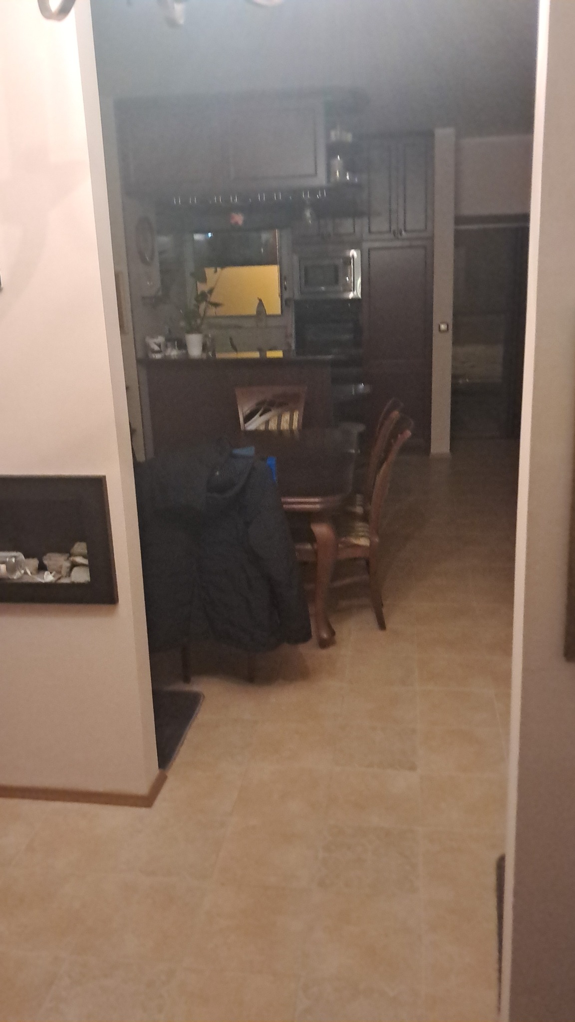 Ujęcie z perspektywy wejścia do salonu z widoczną kuchnią w tle. Na pierwszym planie fragment ściany z kominkiem i krzesła przy stole jadalnym.