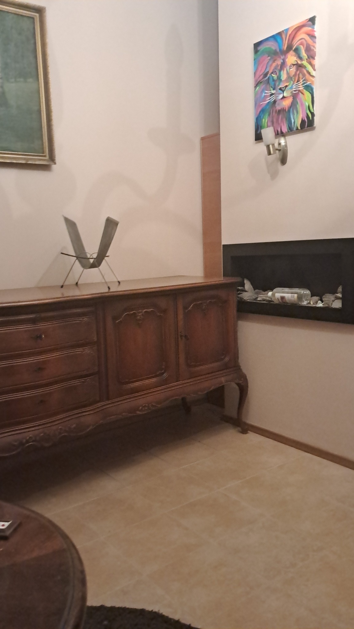 Antyczny kredens z dekoracyjnymi rzeźbieniami, na tle ściany z obrazem lwa i nowoczesnym kominkiem. Widoczna podłoga z płytek w ciepłym odcieniu.