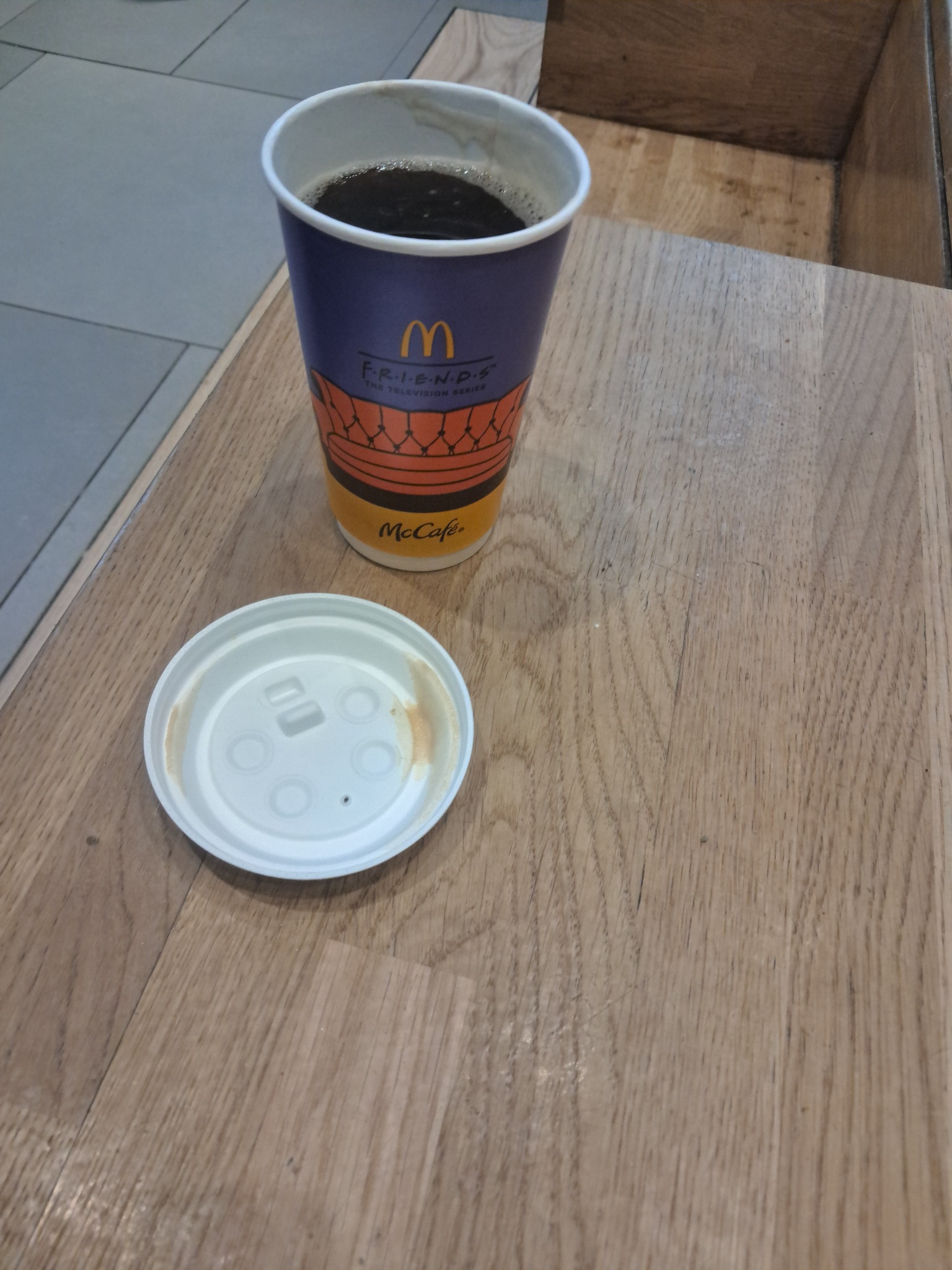 Kawa McCafé w papierowym kubku z motywem Friends, obok biała pokrywka, na drewnianym blacie. Widok z góry, oświetlenie naturalne.