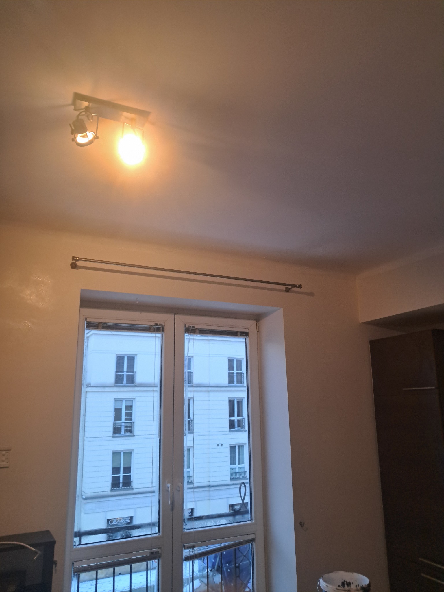 Ściana po gładzi z widocznym oknem i karniszem, oświetlona lampą sufitową. Widoczny fragment szafy i wiaderko po farbie. Ściana w kolorze złamanej bieli.