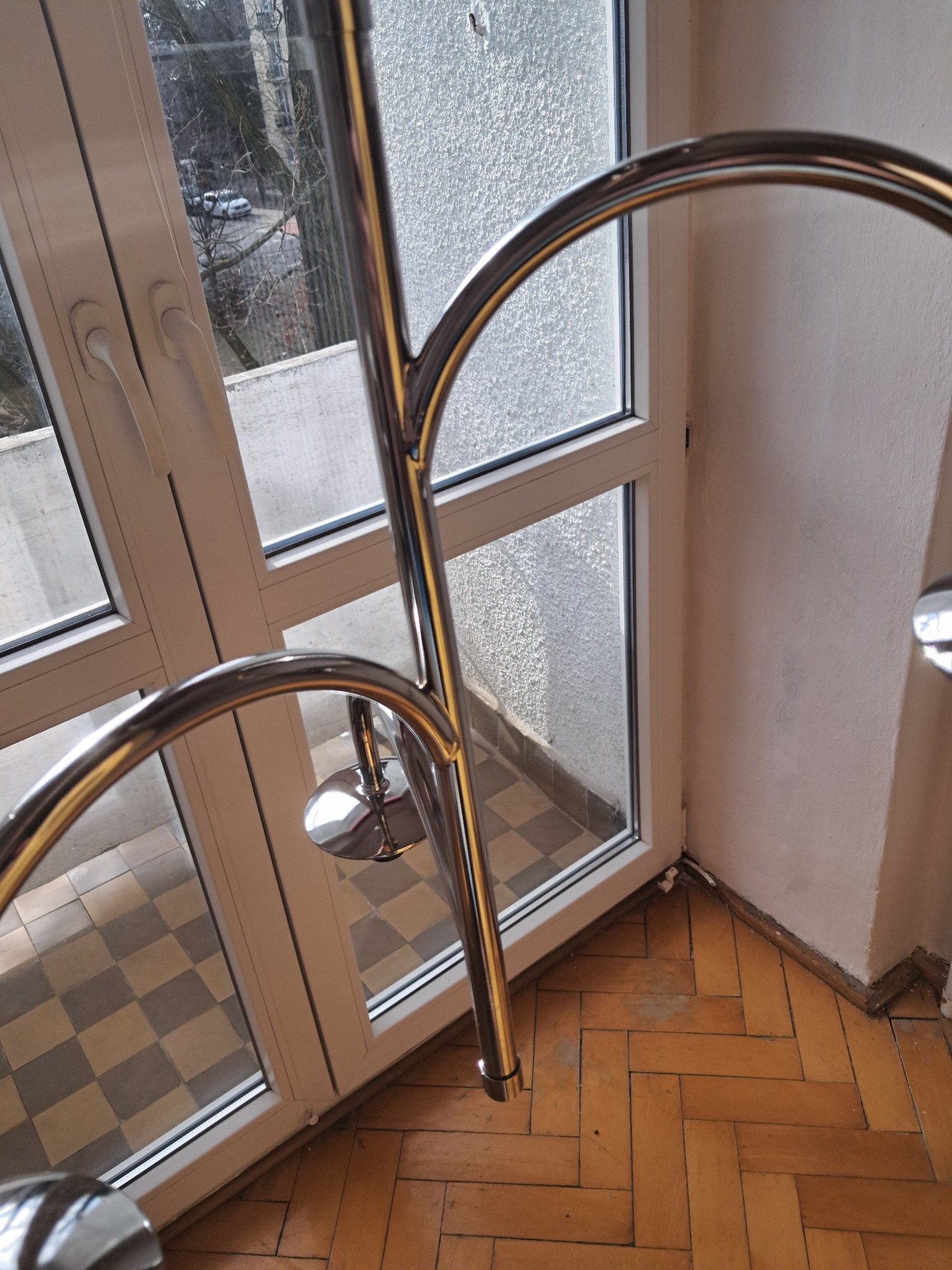 Fragment balkonu z parkietem w jodełkę i metalową barierką, widok z góry. Okno balkonowe z widokiem na zewnątrz. Częściowo widoczna ściana z nierówną fakturą.