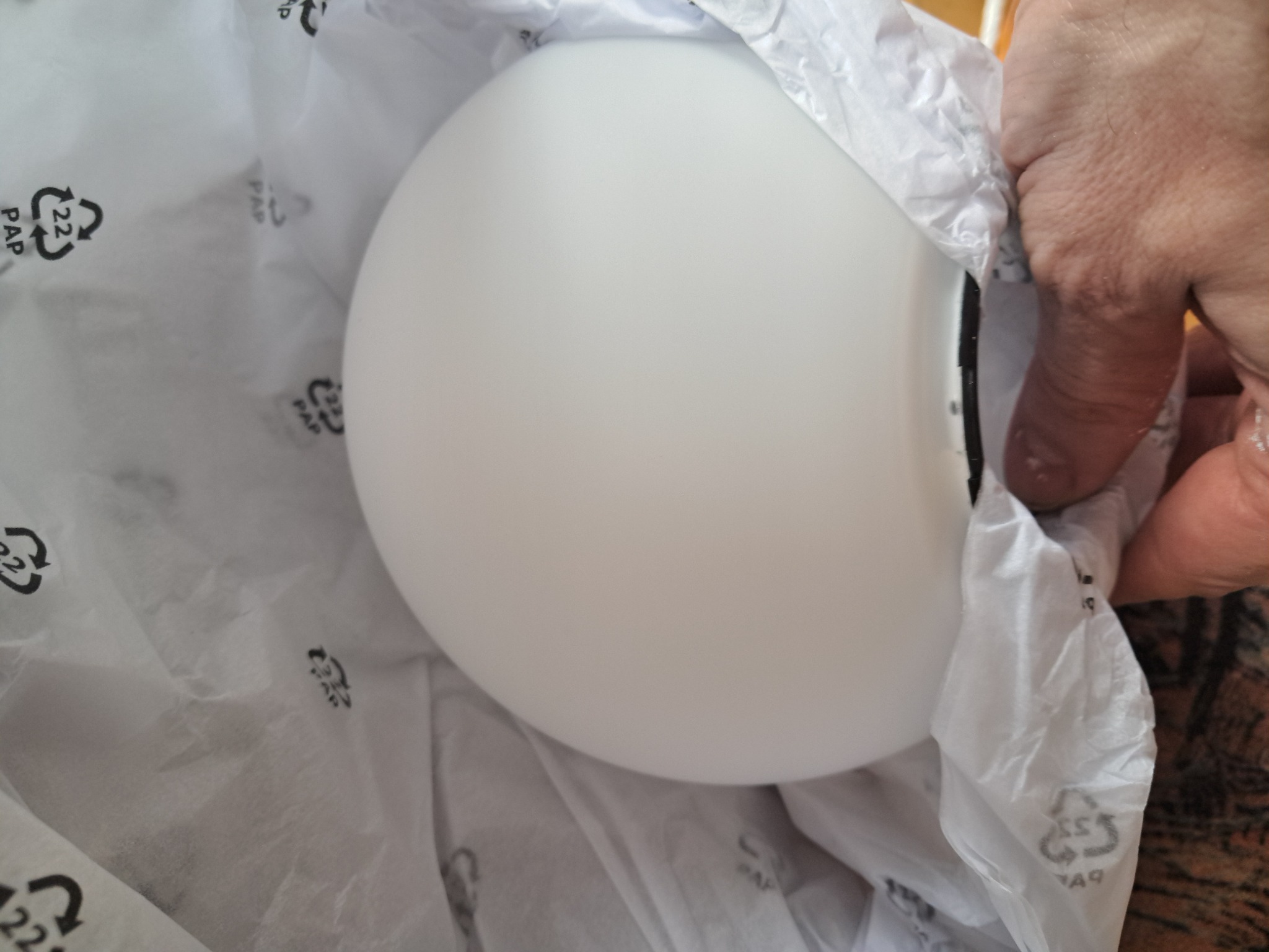 Biała, okrągła lampa w papierowym opakowaniu, częściowo wyjęta z pudełka. Widoczna dłoń osoby wyjmującej lampę. Delikatne światło podkreśla gładką powierzchnię.