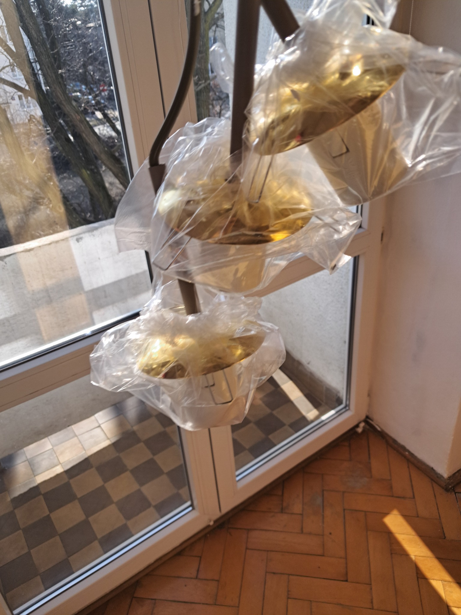 Nowa, złota lampa wisząca, jeszcze w folii ochronnej, w rogu pokoju z parkietem i oknem. Widok na zewnątrz przez okno.