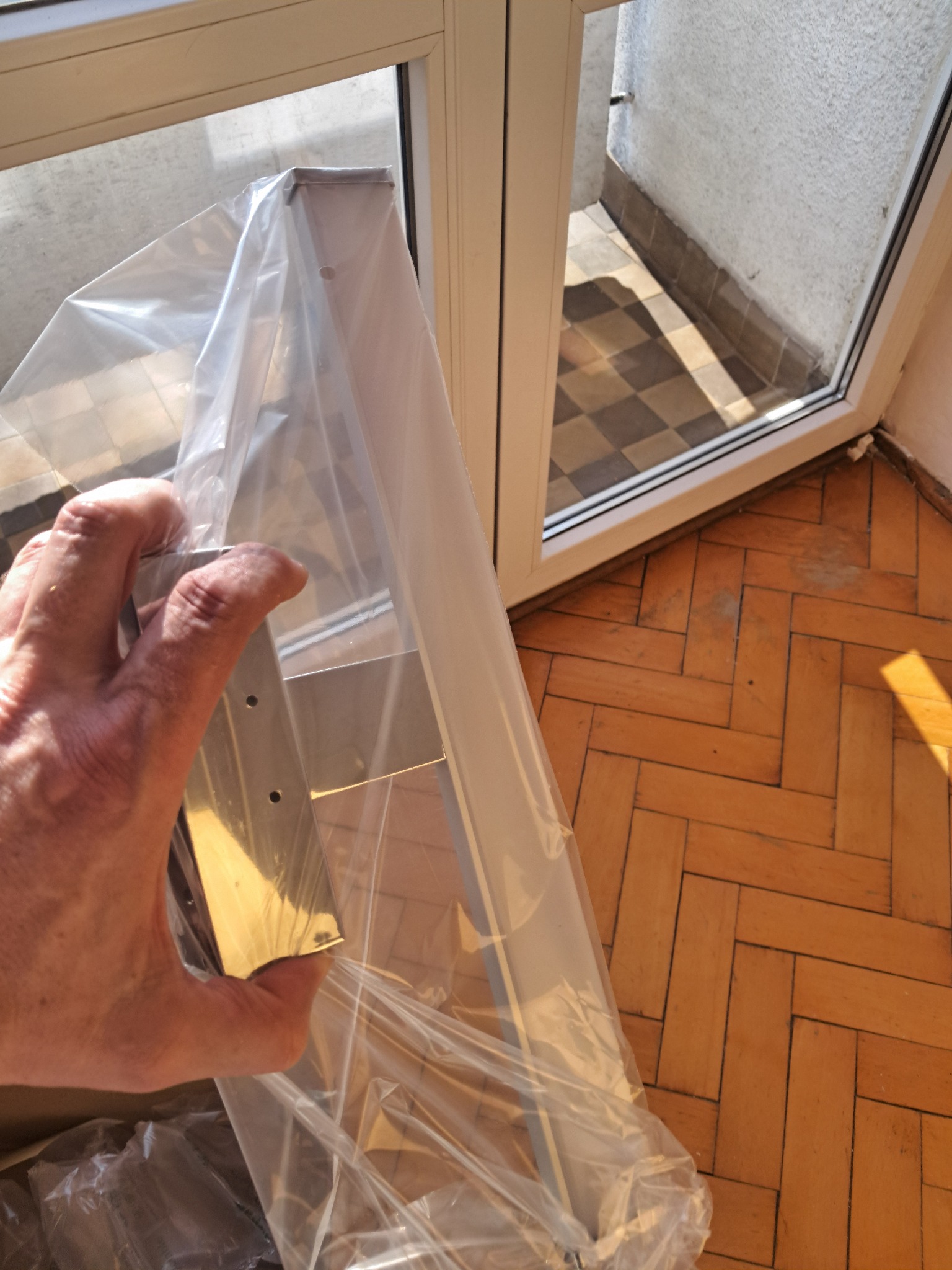 Nowe, srebrne okucie okienne w folii ochronnej, trzymane w dłoni na tle parkietu i okna balkonowego. Przygotowanie do montażu.