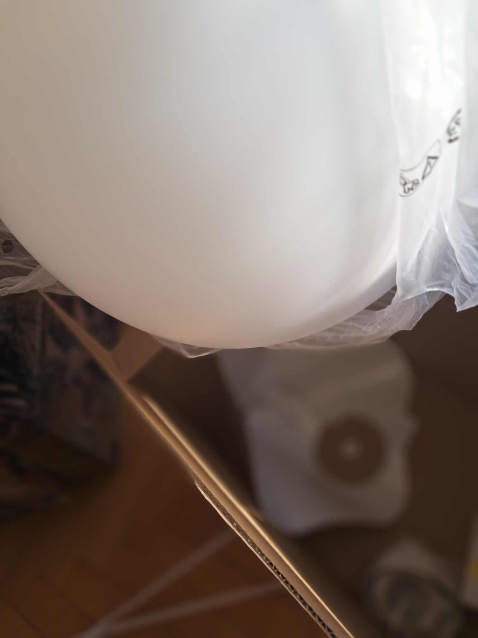 Nowa, biała lampa sufitowa w folii ochronnej, częściowo wyjęta z kartonowego pudełka na drewnianej podłodze. Przygotowanie do montażu oświetlenia.