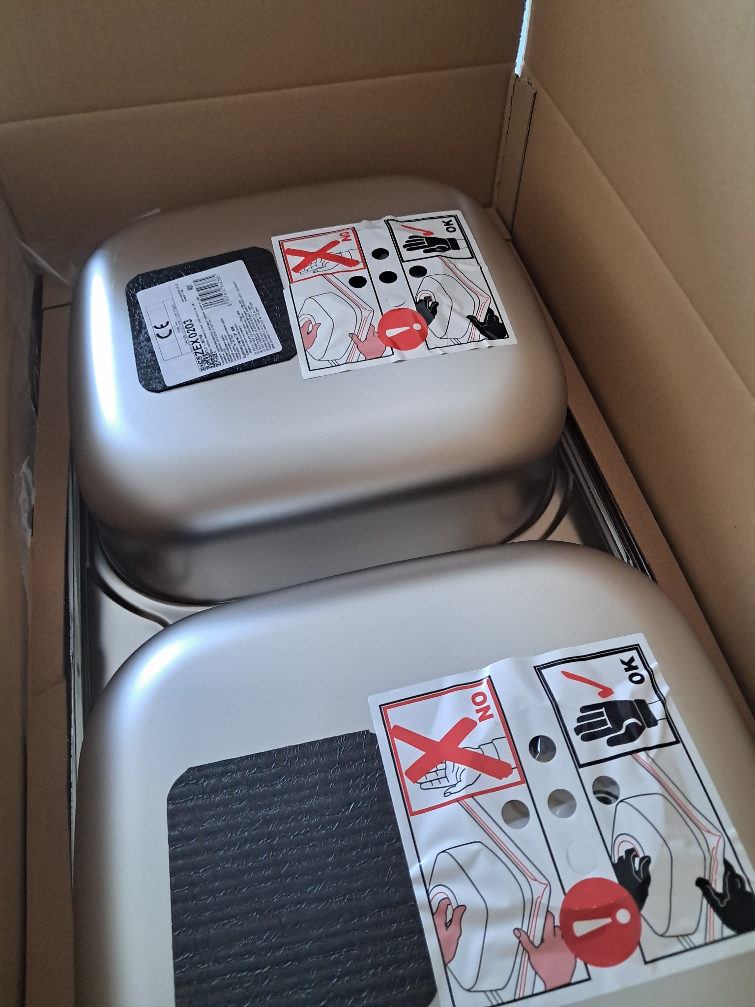 Dwa nowe, nierdzewne zlewozmywaki w kartonowym opakowaniu, z naklejkami instruktażowymi. Widok z góry, oświetlenie naturalne.