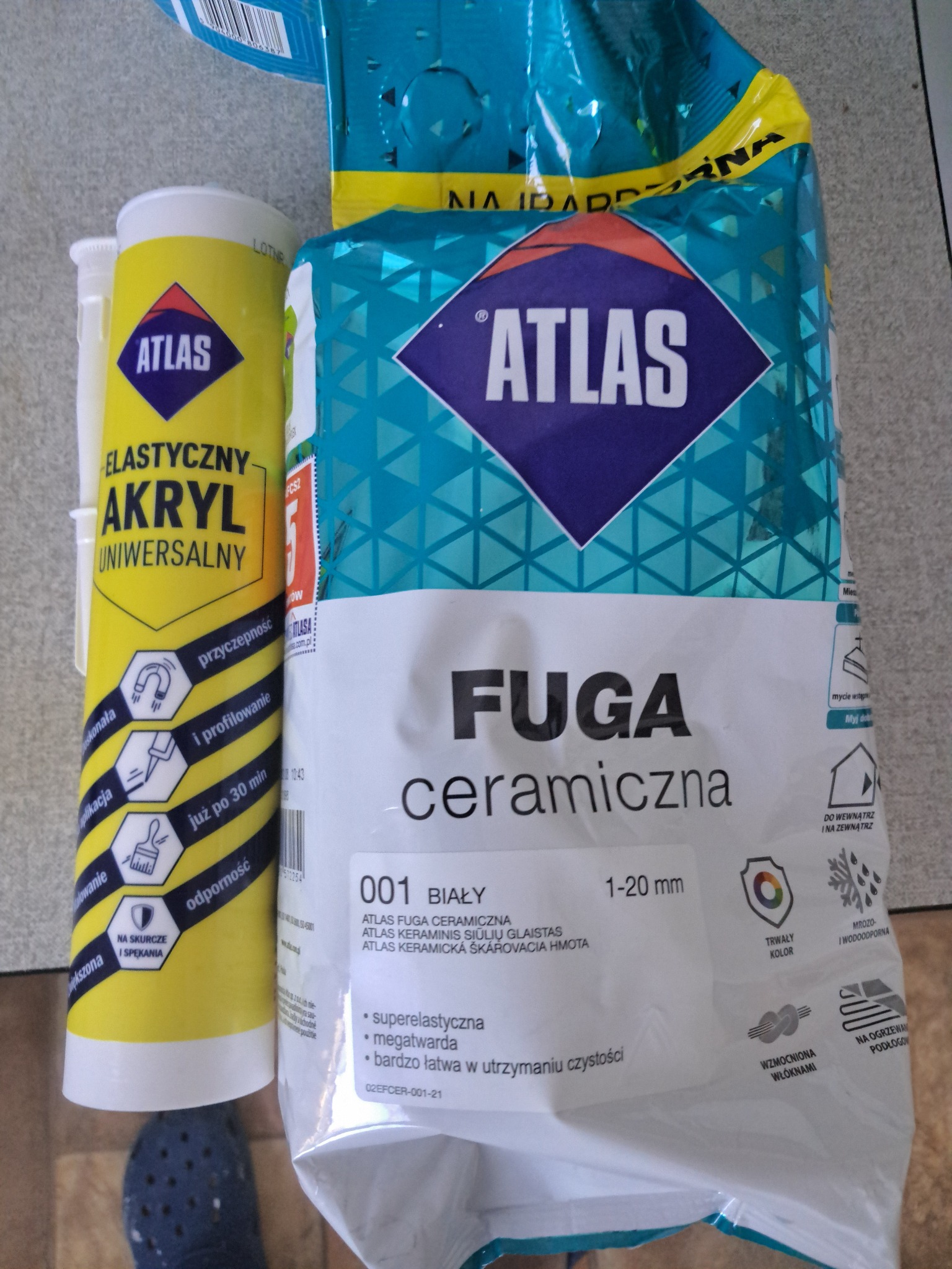 Akryl uniwersalny Atlas i biała fuga ceramiczna Atlas w opakowaniu. Produkty do wykończenia wnętrz, widoczny fragment niebieskiego buta.