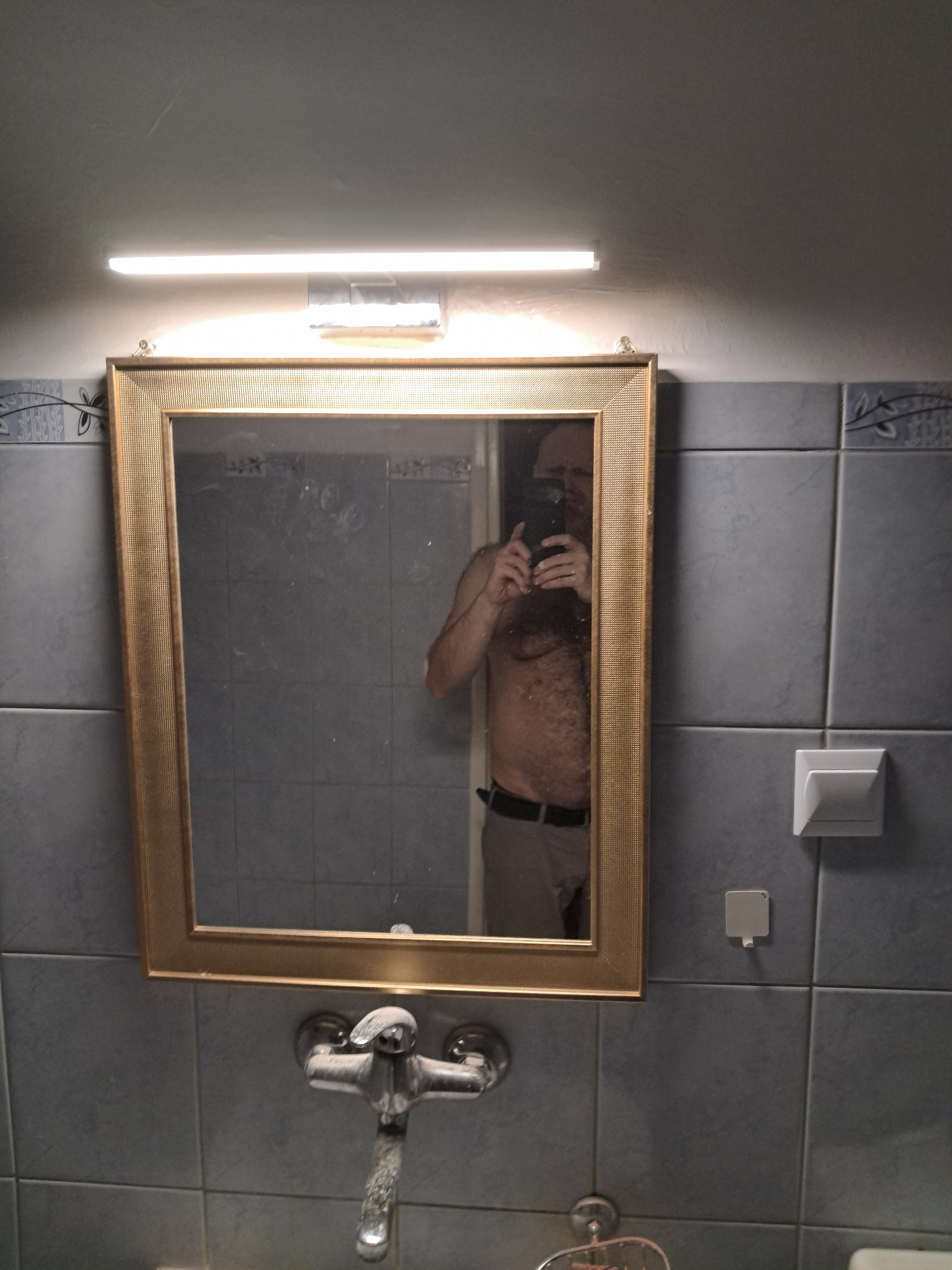 Łazienka z szarymi płytkami i złotym lustrem, w którym odbija się osoba robiąca zdjęcie telefonem. Widoczna armatura i oświetlenie nad lustrem.