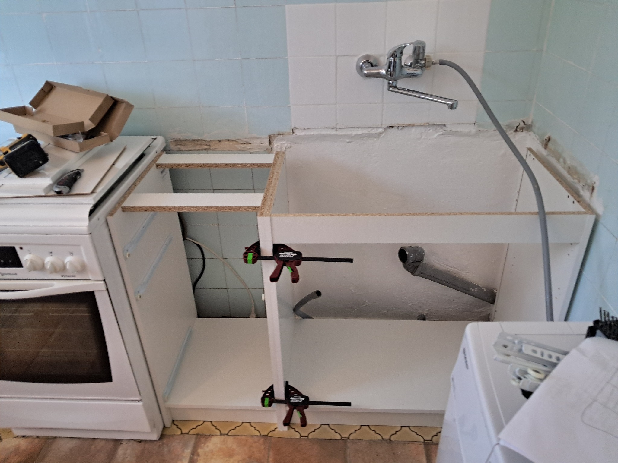 Montaż szafki pod zlew w kuchni. Widoczne klamry stolarskie, fragment kuchenki i pralki. Białe fronty szafek, niebieskie płytki na ścianie.