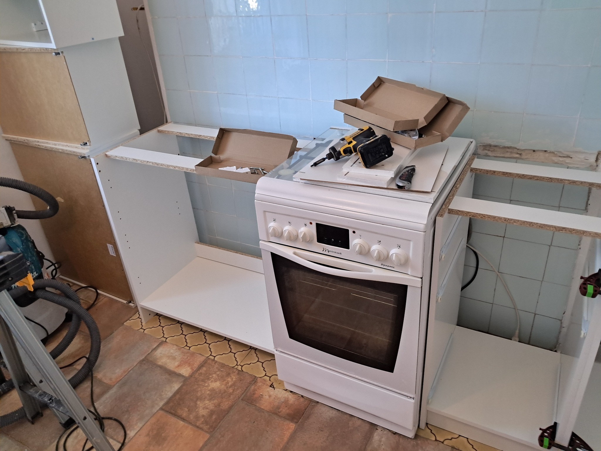 Montaż białych szafek kuchennych w trakcie realizacji. Na kuchence narzędzia i kartony. Widoczne płytki na ścianie i podłoga. W tle odkurzacz.
