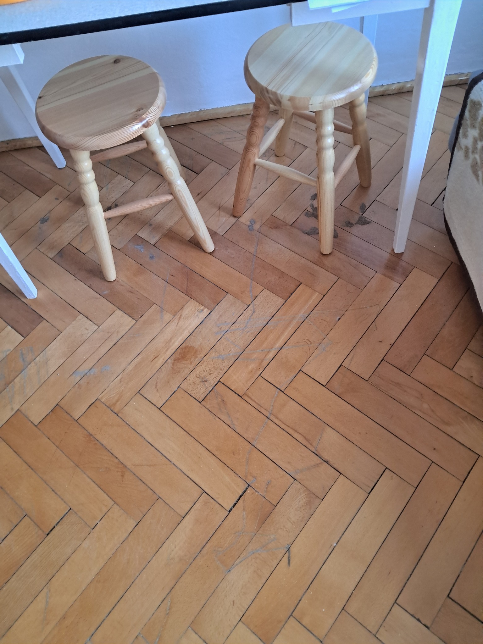 Parkiet ułożony w jodełkę, widoczne dwa drewniane stołki i fragment białego stołu. Podłoga z widocznymi śladami użytkowania, rysami i zabrudzeniami.